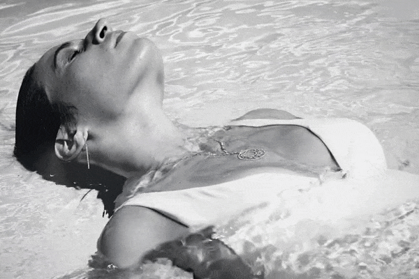 Weibliches Model im Badeanzug, im Pool auf dem Rücken treibend, mit einer Diamantkette und Diamantohrringen.