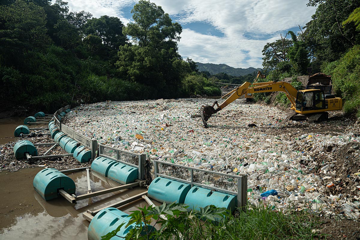 Ein Fluss mit einer Barrikade aus schwimmenden Tonnen, die abertausende weiße Plastikflaschen zurückhält, und ein Bagger, der den Müll aufgreift.