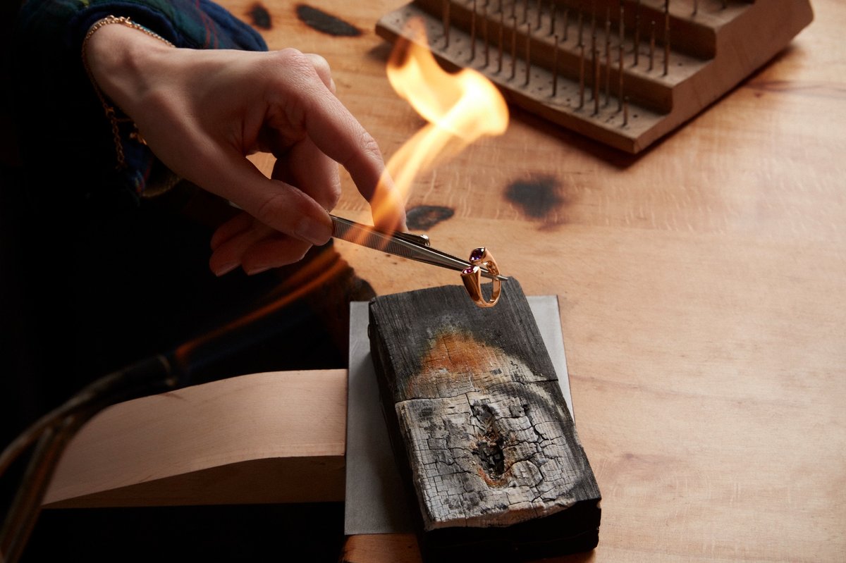 Symbolbild: Eine Hand hält einen Ring mit einer Zange neben die Flamme einer Lötlampe über einem verkohlten Holzblock.