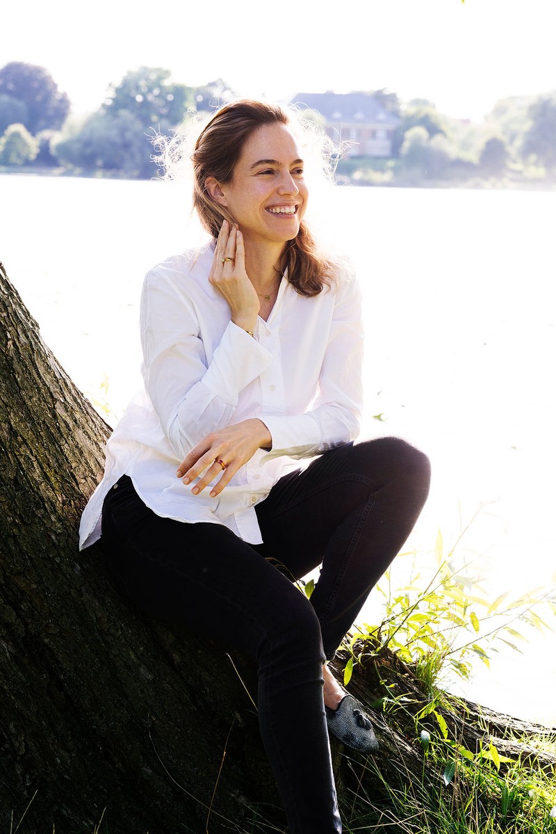 Porträt von Schmuckdesignerin Desiree Sielaff im Freien, sitzend an einem Baum am Wasser, in weißem Hemd und mit Ringen aus der eigenen Kollektion an den Händen.