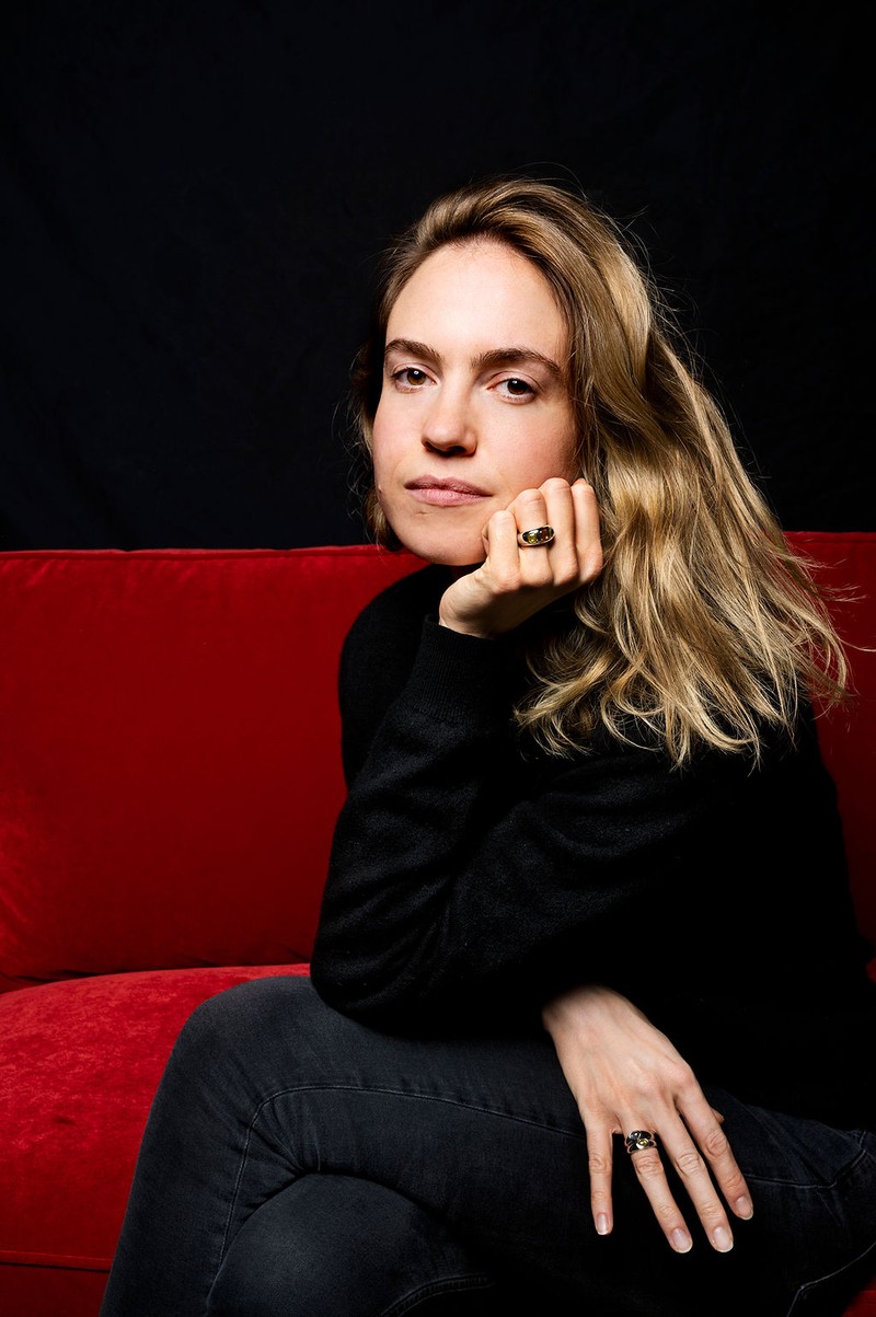 Porträt von Schmuckdesignerin Desiree Sielaff, sitzend auf einem roten Sofa, mit schwarzen Kleidungsstücken und goldenen Ringen mit Farbsteinen.