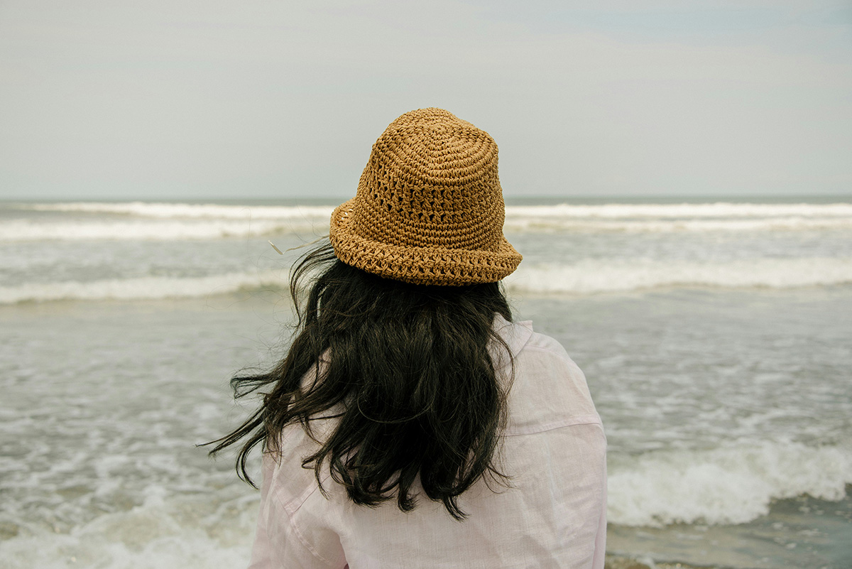 Rückenansicht: Frau mit Strohhut und weißer Bluse schaut aufs Meer – echter UV-Schutz gelingt meist nur mit spezieller Kleidung.