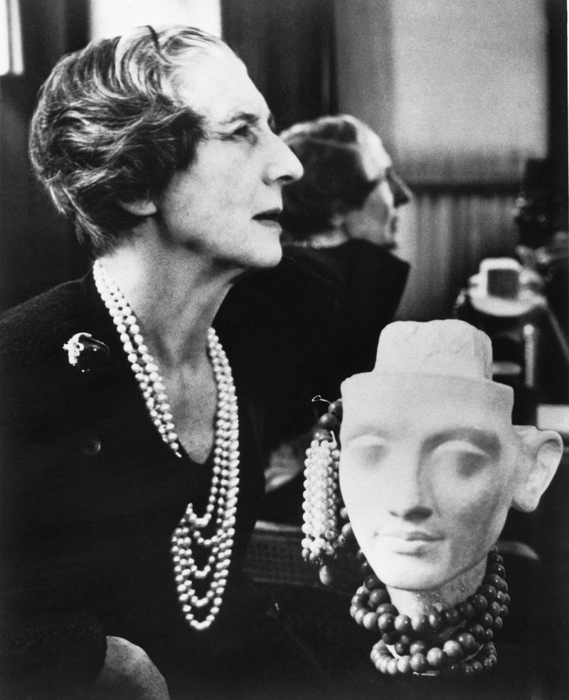 Porträtfotografie: Jeanne Toussaint, Kreativdirektorin von Cartier ab 1933, sitzt im eleganten Kleid mit Perlenkette vor einer Kopfskulptur und einem Spiegel; Schwarzweißaufnahme aus dem Cecil Beaton Archive.