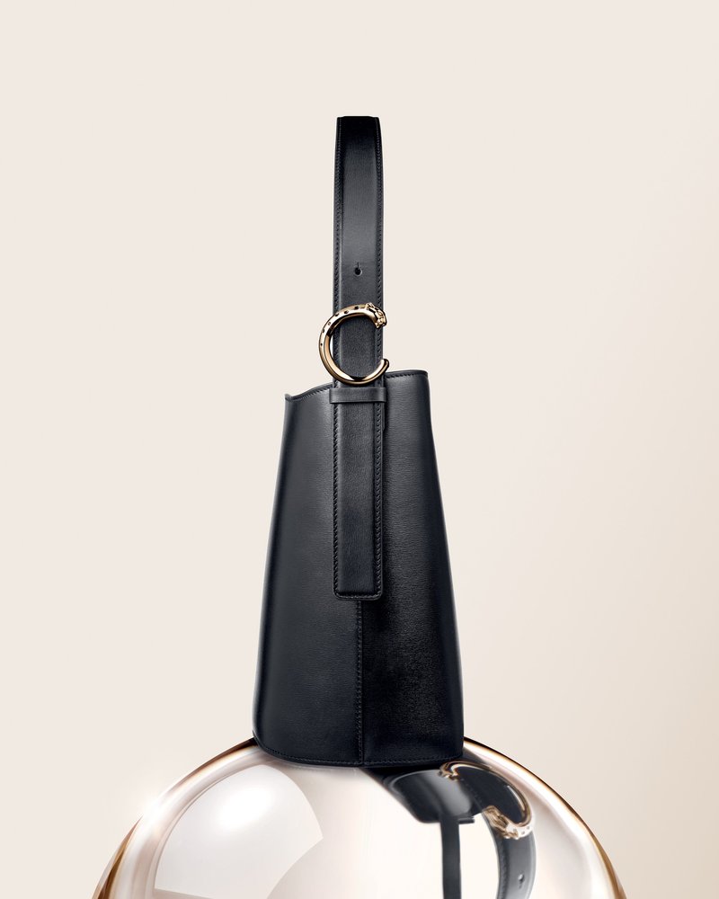 Produktfoto: Panthère C de Cartier Bucket Bag in schwarzem Kalbsleder, seitliche Ansicht; goldfarbene Panther-Schnalle am Riemen, Tasche ruht auf einer glänzenden Kugel.