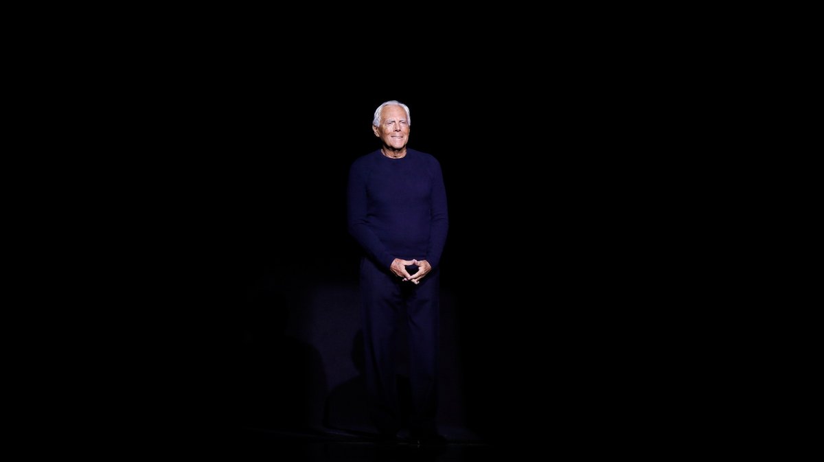Giorgio Armani steht allein im Scheinwerferlicht auf einer dunklen Bühne.
