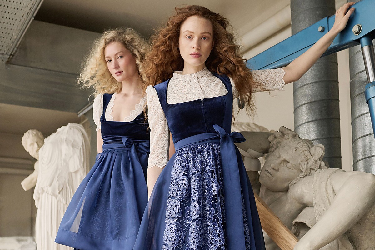 Zwei Models in marineblauen Samtdirndln stehen in einem Atelier mit Skulpturen, links ein kürzeres Dirndl mit transparenter Spitzenbluse, rechts ein Midi-Dirndl mit floraler Spitzen-Schürze, beide mit seitlich gebundenen Schleifen.