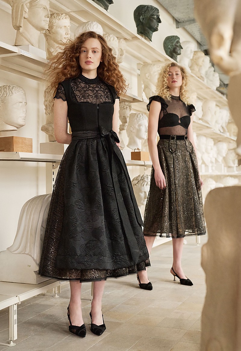 Zwei Models in schwarzen Abend-Dirndl-Looks stehen in einem Saal mit Skulpturen, links ein Midi-Dirndl mit Schnürung und transparenter Spitzenbluse, rechts ein glitzernder Midirock mit transparentem, gepunktetem Body, beide mit Absatzschuhen.