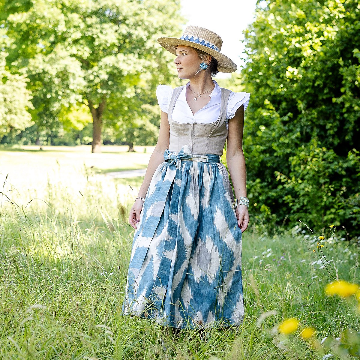 Frau steht in einer sommerlichen Wiese, trägt ein helles Leinen-Dirndl mit weißer Bluse und eine blau weiße Ikat-Schürze, seitlich gebundene Schleife, Strohhut mit Band, Bäume im Hintergrund, Tageslicht.