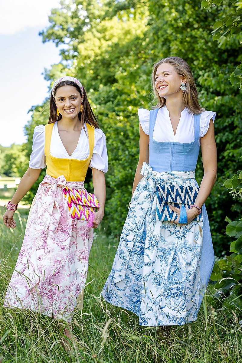 Zwei Frauen gehen lachend durch eine Wiese, beide tragen Dirndl mit weißer Bluse, eine in Gelb mit rosa gemusterter Schürze, die andere in Hellblau mit blauer Schürze, jeweils mit seitlich gebundener Schleife, Bäume im Hintergrund, Tageslicht.
