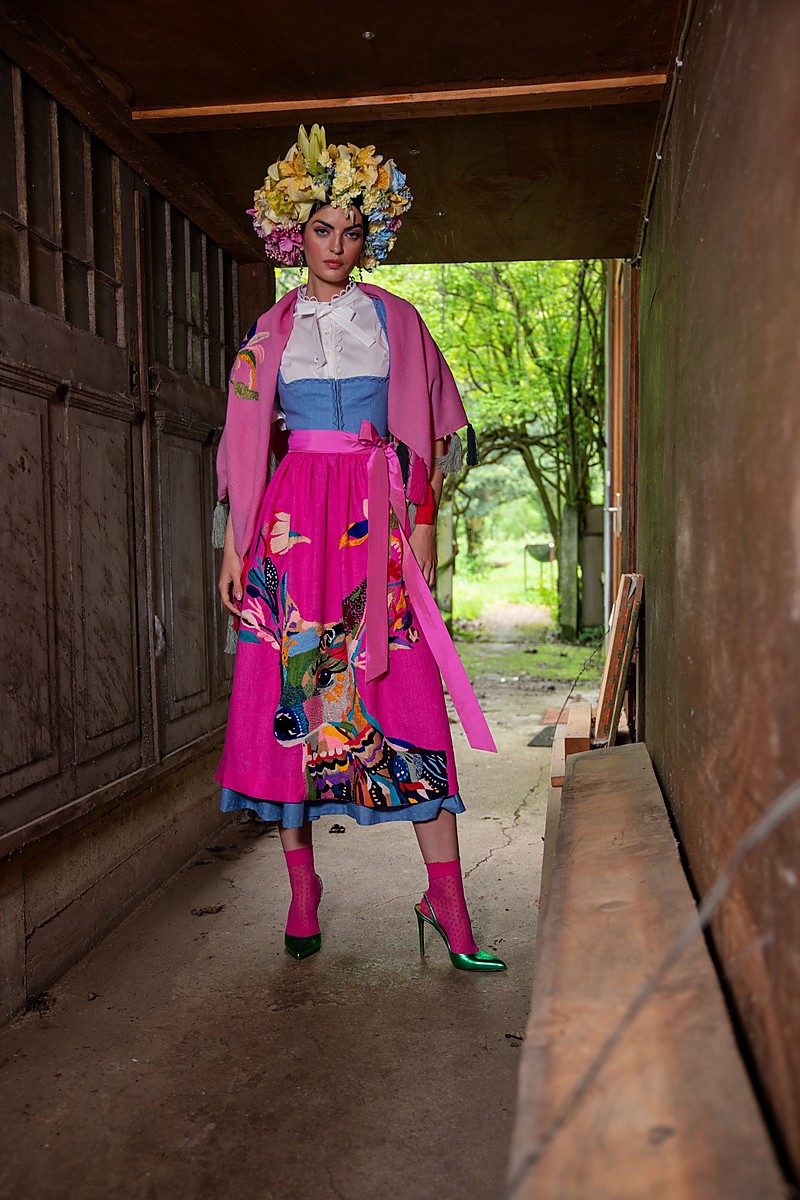 Modell steht in einem überdachten Durchgang, trägt ein blaues Dirndl mit rosa Schürze und passendem Tuch, auf der Schürze ein großes, buntes Hirschmotiv, darüber offene Umgebung mit Bäumen im Hintergrund.