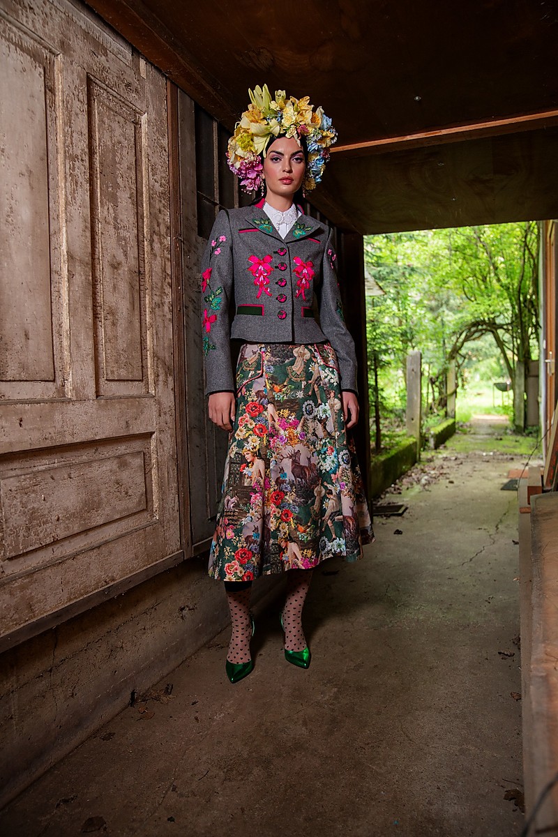 Modell steht in einem überdachten Durchgang, trägt eine graue, kurz geschnittene Trachtenjacke mit kräftigen floralen Stickereien und farbigen Paspeln, dazu ein weit schwingender Brokatrock mit vielfarbigem Motiv, grüne Pumps und gepunktete Strümpfe, Bäume im Hintergrund.
