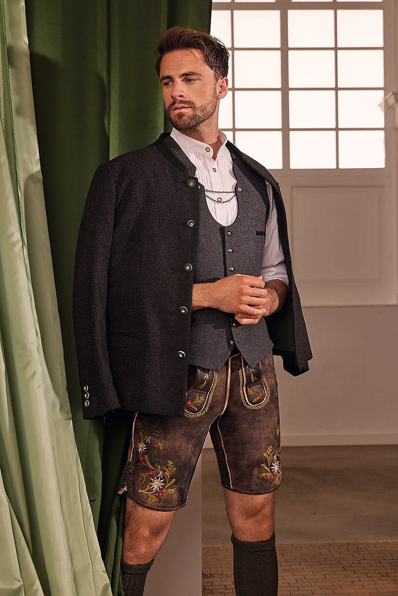 Mann in klassischer Tracht, naturbraune kurze Lederhose, weißes Hemd und dunkle Trachtenjacke, knielange Strümpfe und Haferlschuhe, steht frontal zur Kamera.