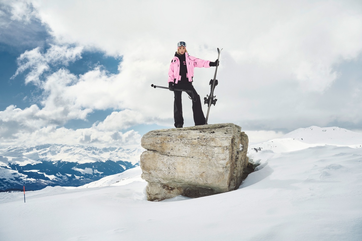 Person in pinker Elho-Bomberjacke steht mit Skiern auf einem Felsen im Schnee, Alpenpanorama im Hintergrund