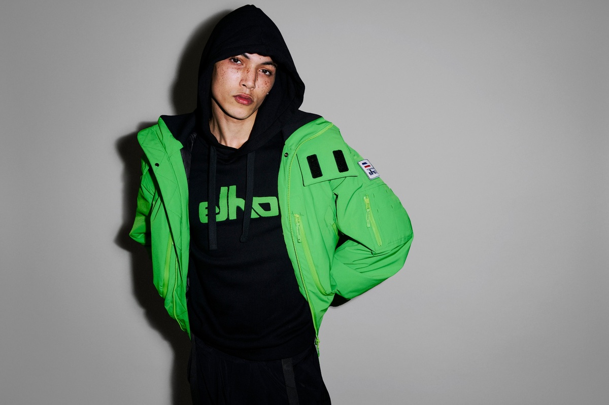 Person trägt eine grüne Elho-Bomberjacke über schwarzem Hoodie, Studioaufnahme vor grauem Hintergrund