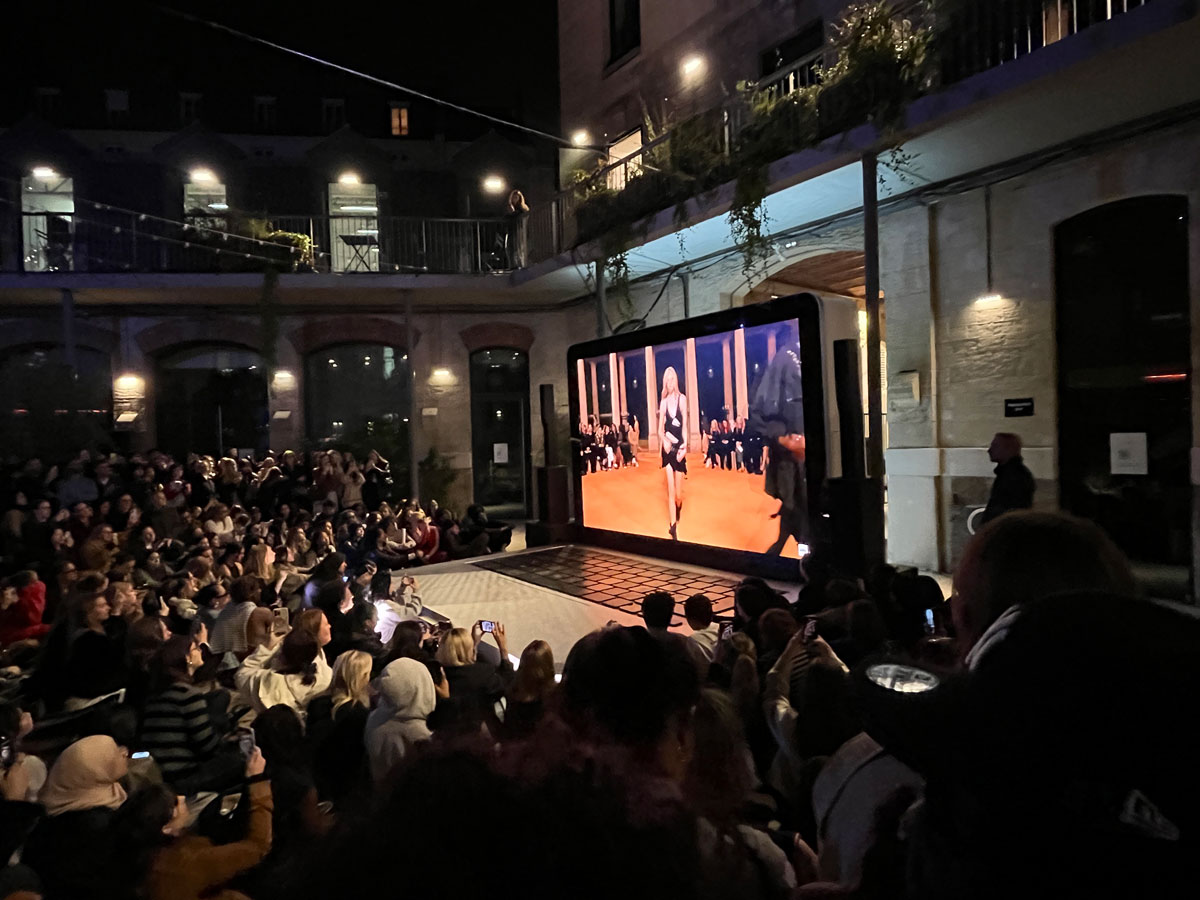Open-Air-Veranstaltung in einem Pariser Innenhof: Das Defilee läuft auf einem bühnengroßen Laptop, davor sitzen zahlreiche Leute und verfolgen die Show im Public Viewing.