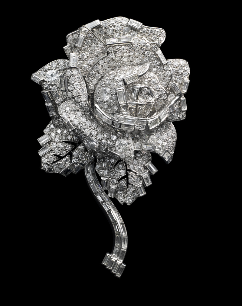 substantial-times-cartier-im-victoria-and-albert-museum-glanz-macht-und-mythos-rosenbrosche-08 Rosettenförmige Clip-Brosche von Cartier London (1938) aus Diamanten und Platin; plastisch modellierte Blütenblätter und gebogener Stiel, freigestellt vor schwarzem Hintergrund.