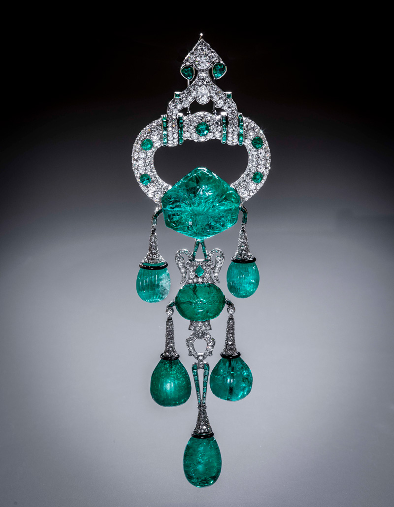 substantial-times-cartier-im-victoria-and-albert-museum-glanz-macht-und-mythos-smaragdbrosche-Marjorie-Merriweather-Post-04 Hochformatiges Foto einer großen Cartier-Smaragdbrosche (New York, 1928): Platin, Diamanten und mehrere gravierte Smaragd-Tropfen/-Kugeln; reich verzierte Aufhängung, freigestellt vor grauem Verlauf.