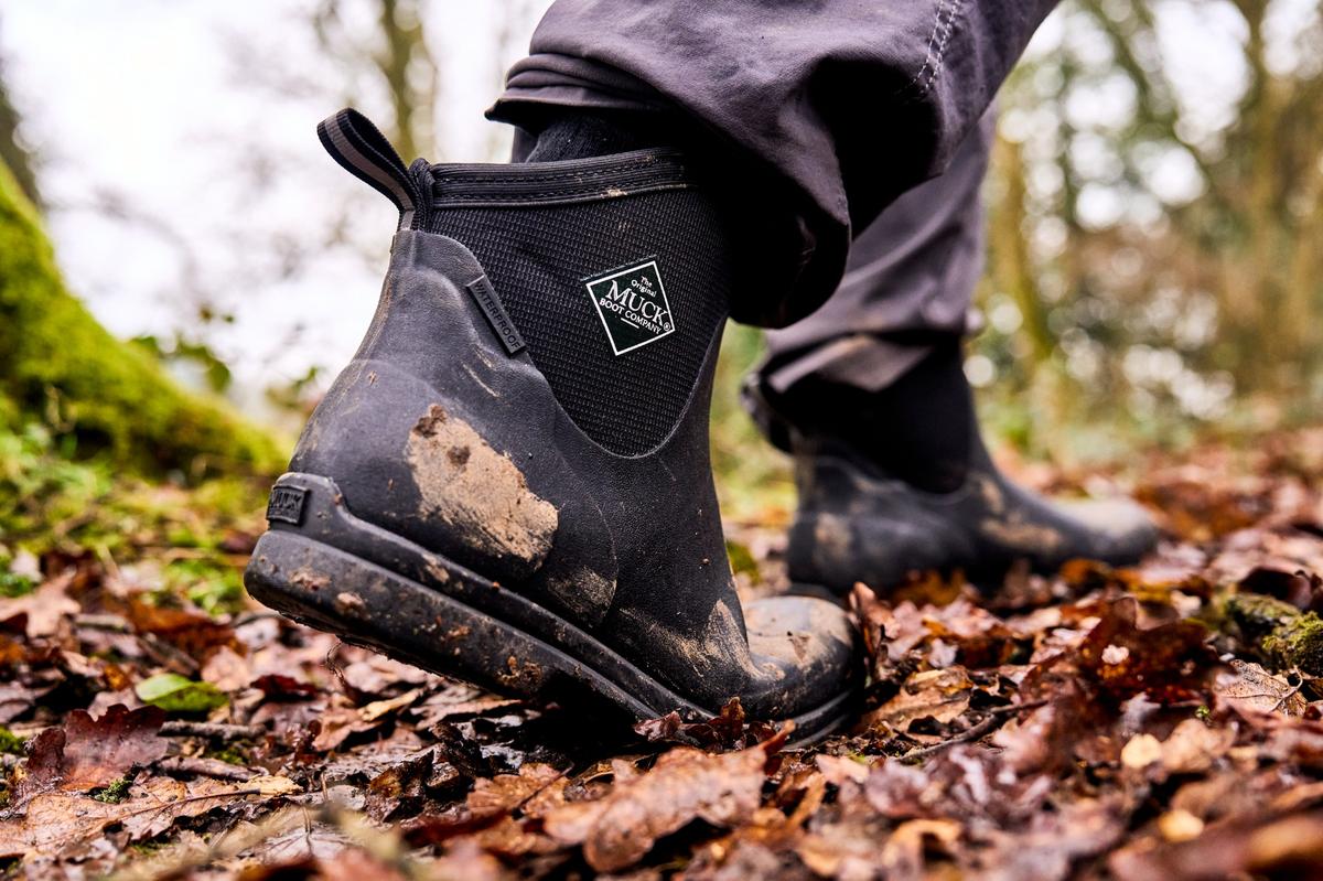 Knöchelhoher schwarzer Outdoor-Boot mit Muck-Logo, von Schlamm und Laub bedeckt, auf Waldboden.