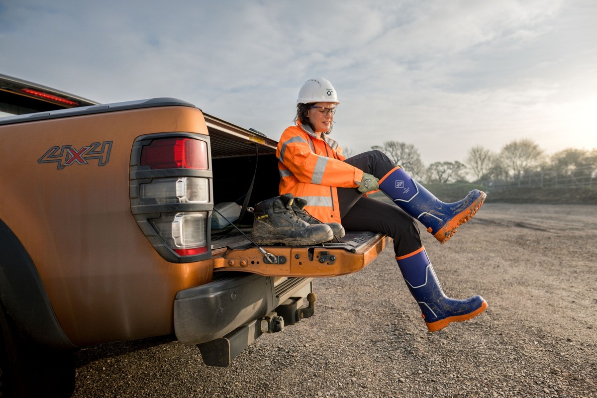 Frau in Arbeitskleidung sitzt auf Laderampe eines Pick-ups, trägt blaue Sicherheits-Gummistiefel. BU: Preis: 159 Euro Foto-Credit: