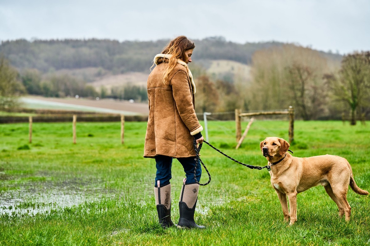 Frau führt Hund auf nasser Wiese, trägt beige-schwarze hohe Gummistiefel.