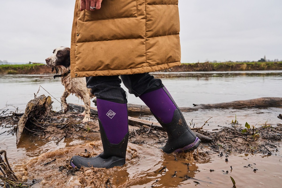 Angeschnittenes Bild: Frau – man sieht nur einen goldgelben Steppmantel von der Taille abwärts – geht mit Hund an schlammigem Flussufer, trägt lilafarbene Muck Boots.