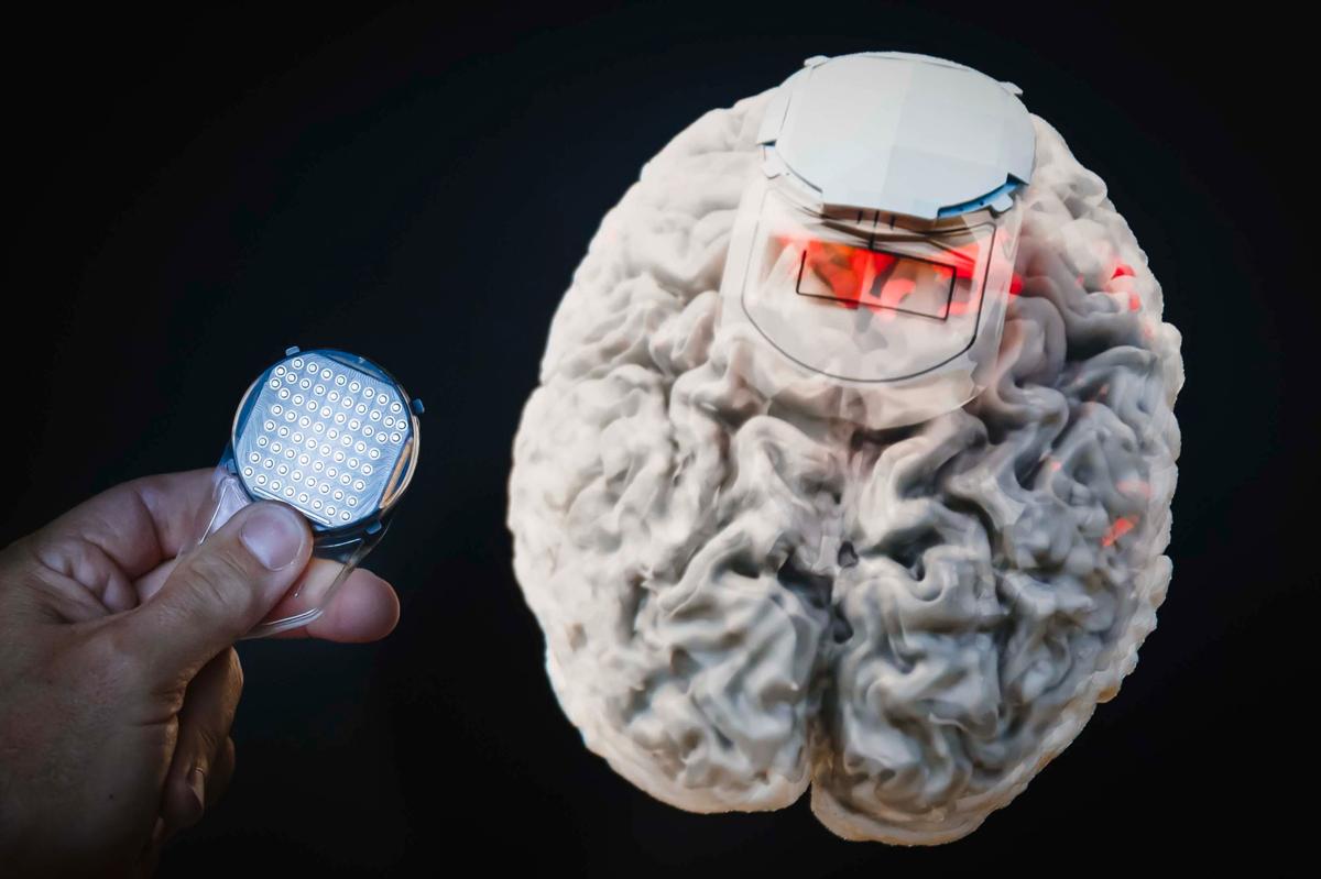 substantial-times-die-bruecke-ueber-das-unmoegliche-gregoire-courtine-rolex-award-01 Detailaufnahme einer Hand, die ein rundes Gehirnimplantat mit zahlreichen Elektrodenflächen hält; daneben ein anatomisches Modell eines Gehirns, in das das Implantat oben eingesetzt ist, teilweise rot beleuchtet zur Darstellung der aktiven Areale.