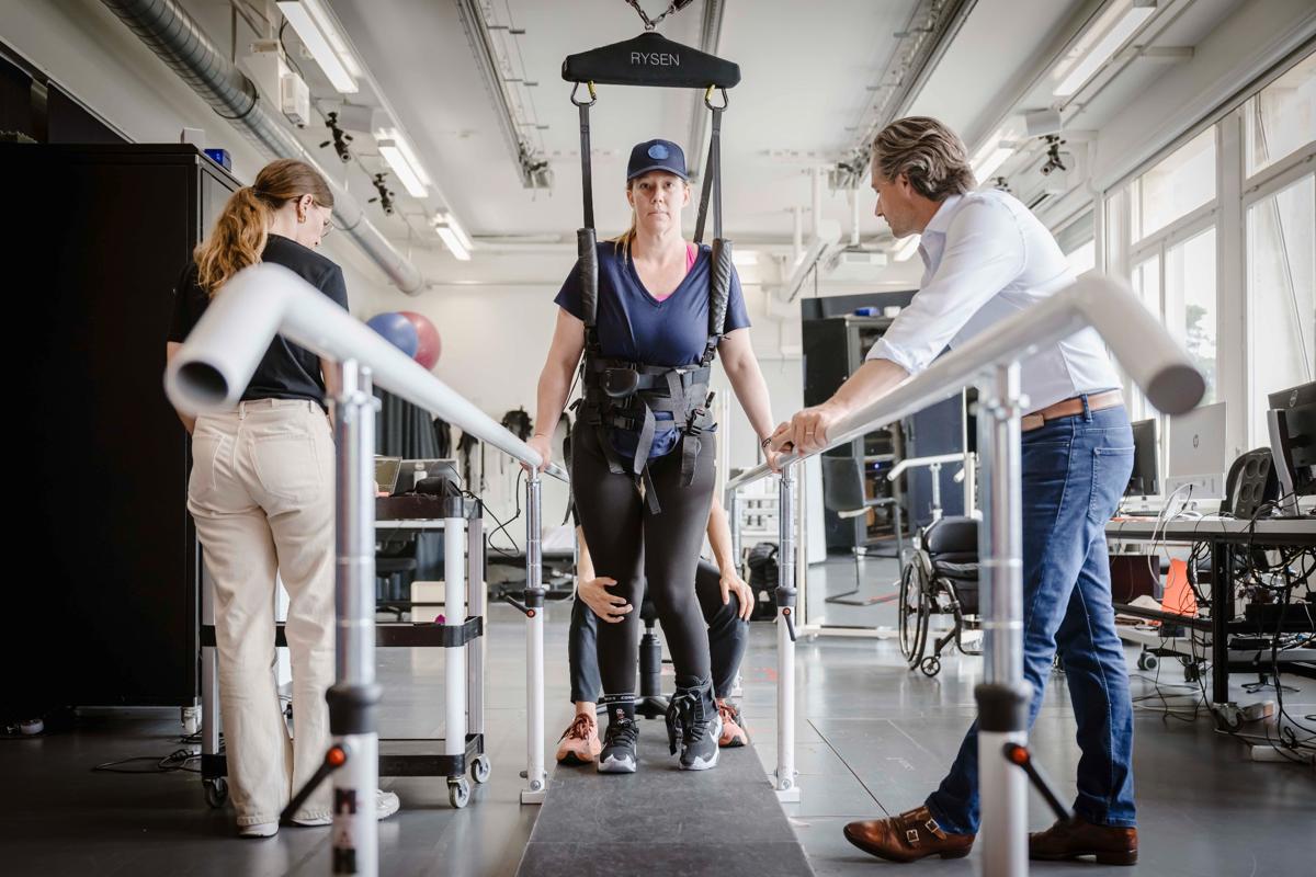 Suzanne Edwards steht in einem Rehabilitationslabor in einem Laufgeschirr, das sie beim Gehen unterstützt; Therapeutinnen sichern und beobachten sie, während Grégoire Courtine am Gehbarren steht und den Ablauf überwacht.