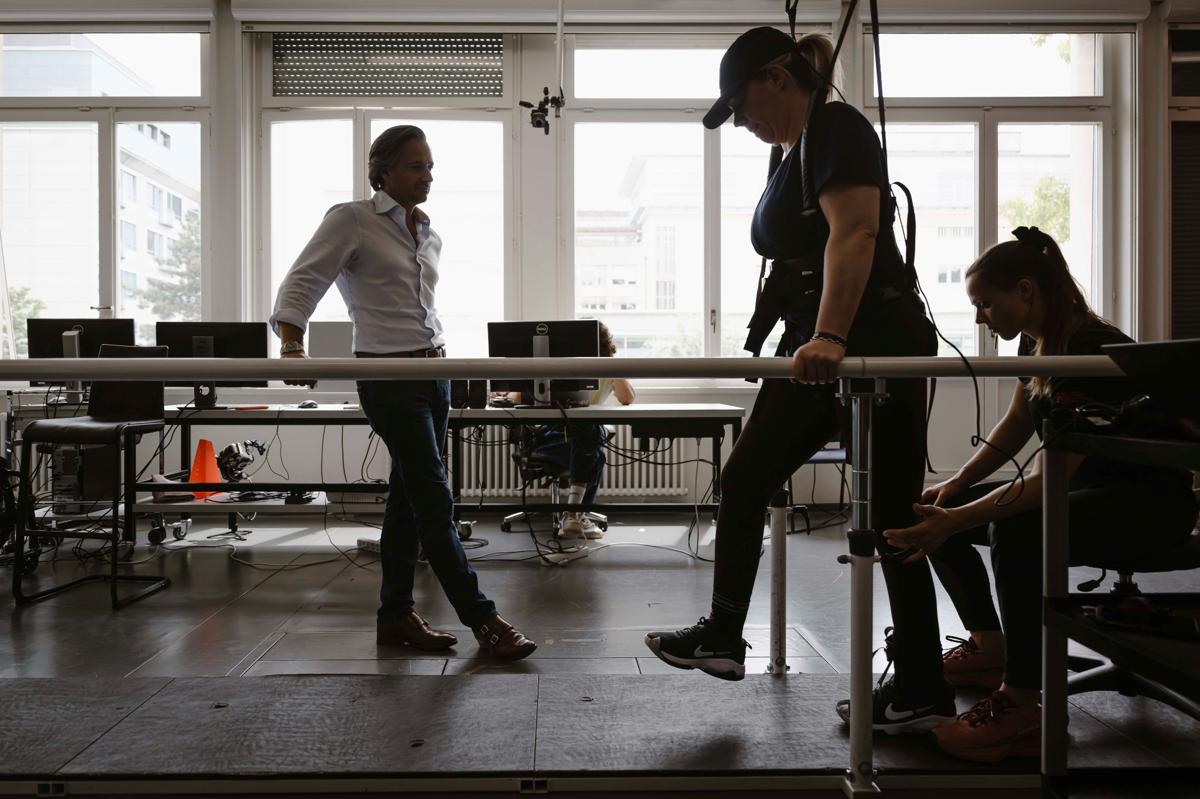 substantial-times-die-bruecke-ueber-das-unmoegliche-gregoire-courtine-rolex-award-03 Suzanne Edwards steht in einem Laufgeschirr an Parallelstützen und bereitet sich auf ihre ersten Schritte vor, während Grégoire Courtine beobachtend danebensteht; eine Therapeutin unterstützt sie an den Beinen.