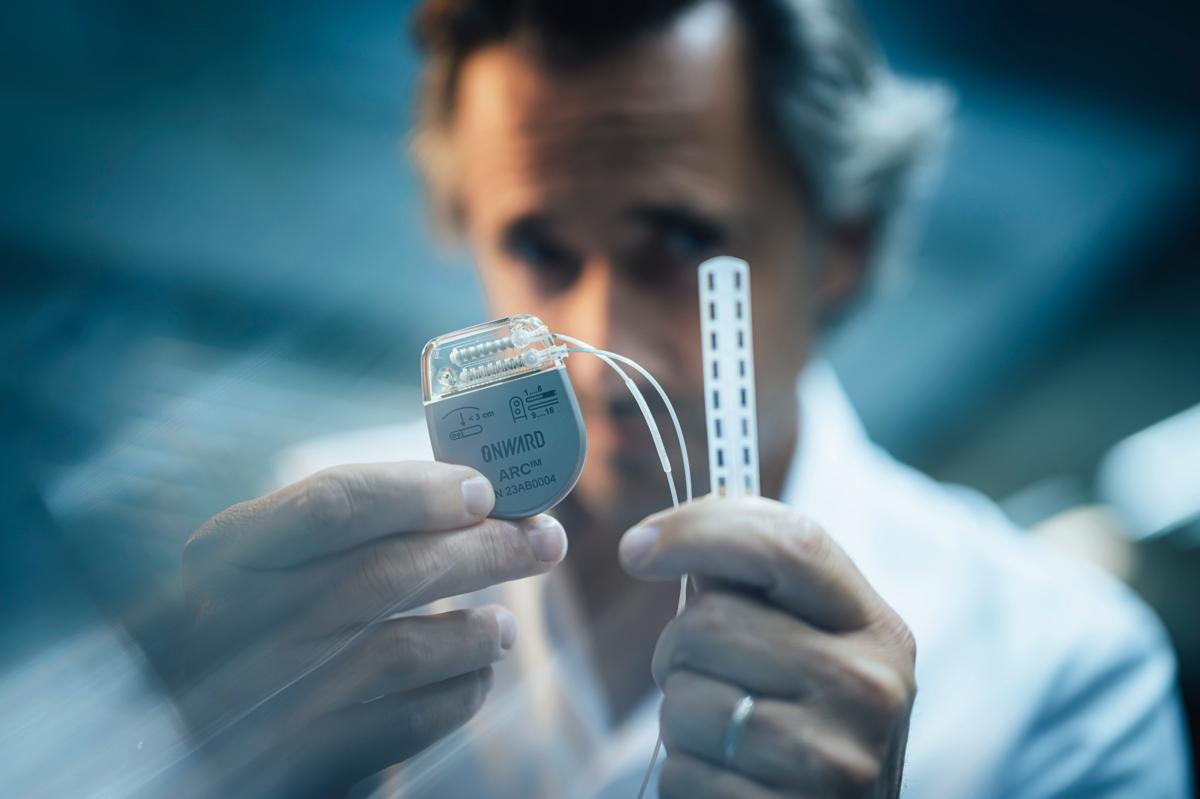 substantial-times-die-bruecke-ueber-das-unmoegliche-gregoire-courtine-rolex-award-05 Grégoire Courtine hält ein implantierbares medizinisches Gerät und eine Elektrodenleiste in den Händen; sein Gesicht ist unscharf im Hintergrund, während das Implantat im Vordergrund deutlich sichtbar ist.