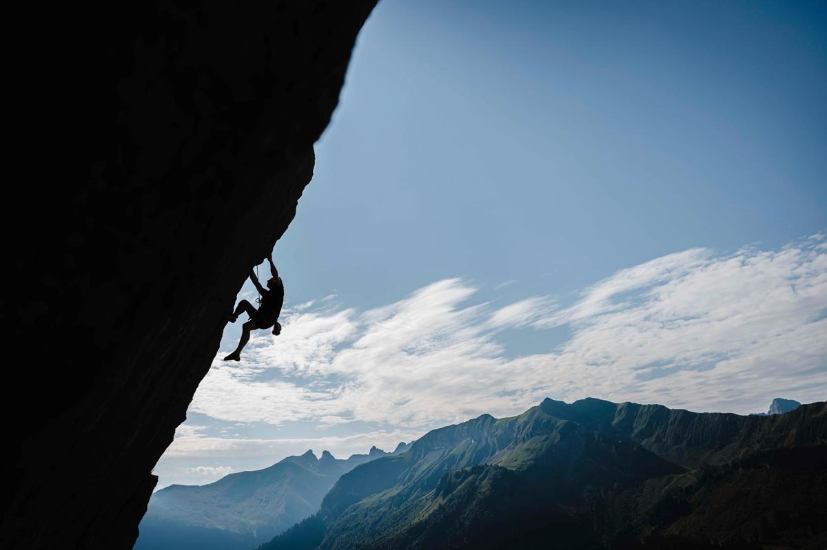 substantial-times-die-bruecke-ueber-das-unmoegliche-gregoire-courtine-rolex-award-08 Silhouette von Grégoire Courtine, der eine steile Felswand in Chamonix erklimmt; unter ihm erstreckt sich ein sonniges Bergpanorama mit klarer Sicht auf Gipfel und Wolken.