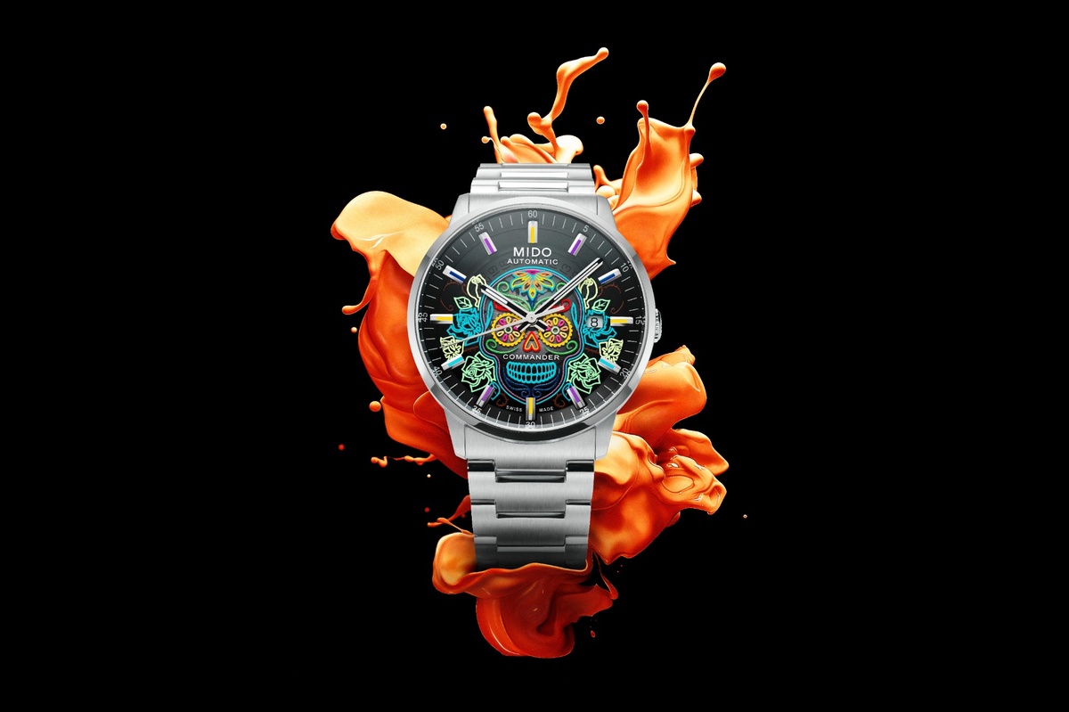 substantial-times-mehr-zeit-als-wir-02 Eine Armbanduhr von Mido mit Edelstahlgehäuse und -armband, deren schwarzes Zifferblatt ein farbenfrohes, leuchtendes Totenkopf-Motiv im Stil des Día de los Muertos zeigt. Hinter der Uhr spritzen orangefarbene Flüssigkeitselemente auf schwarzem Hintergrund, die den lebendigen Charakter des Designs betonen.