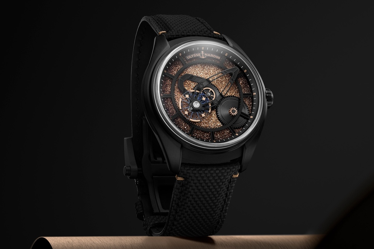 substantial-times-mehr-zeit-als-wir-04 Armbanduhr von Ulysse Nardin, Modell Freak X Crystalium, in einem schwarzen Titangehäuse mit schwarzer Lünette. Das skelettierte Zifferblatt zeigt eine rotierende Stundenscheibe in schimmerndem Roségoldton, deren Struktur an Kristalle erinnert. Sichtbar sind Zahnräder und Brücken in Schwarz sowie blaue Elemente des Uhrwerks. Die Uhr trägt ein schwarzes Kautschukarmband mit textilähnlicher Struktur.