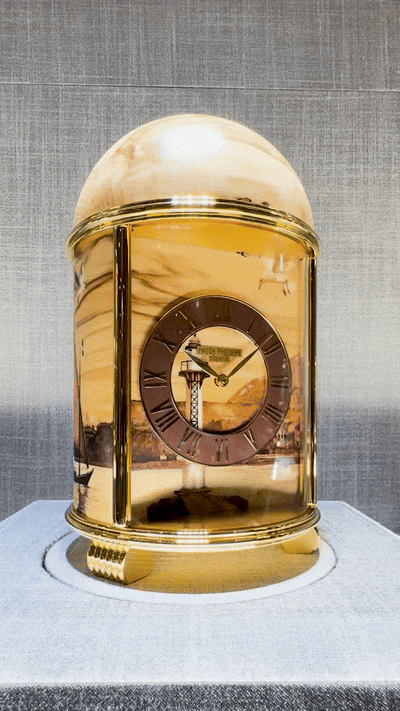 Videoaufnahme einer Kuppel-Tischuhr von Patek Philippe, vollständig in Holzmarqueterie gestaltet. Beim Drehen der Uhr wird das umlaufende Hafenmotiv von Genf sichtbar, einschließlich Jet d’eau, Leuchtturm, Segelbooten und Möwen. Die Kuppel zeigt die natürliche Maserung von gespaltetem Buchenholz. Das Zifferblatt besitzt einen braun lackierten Stundenring mit römischen Ziffern und roségoldenen Zeigern.