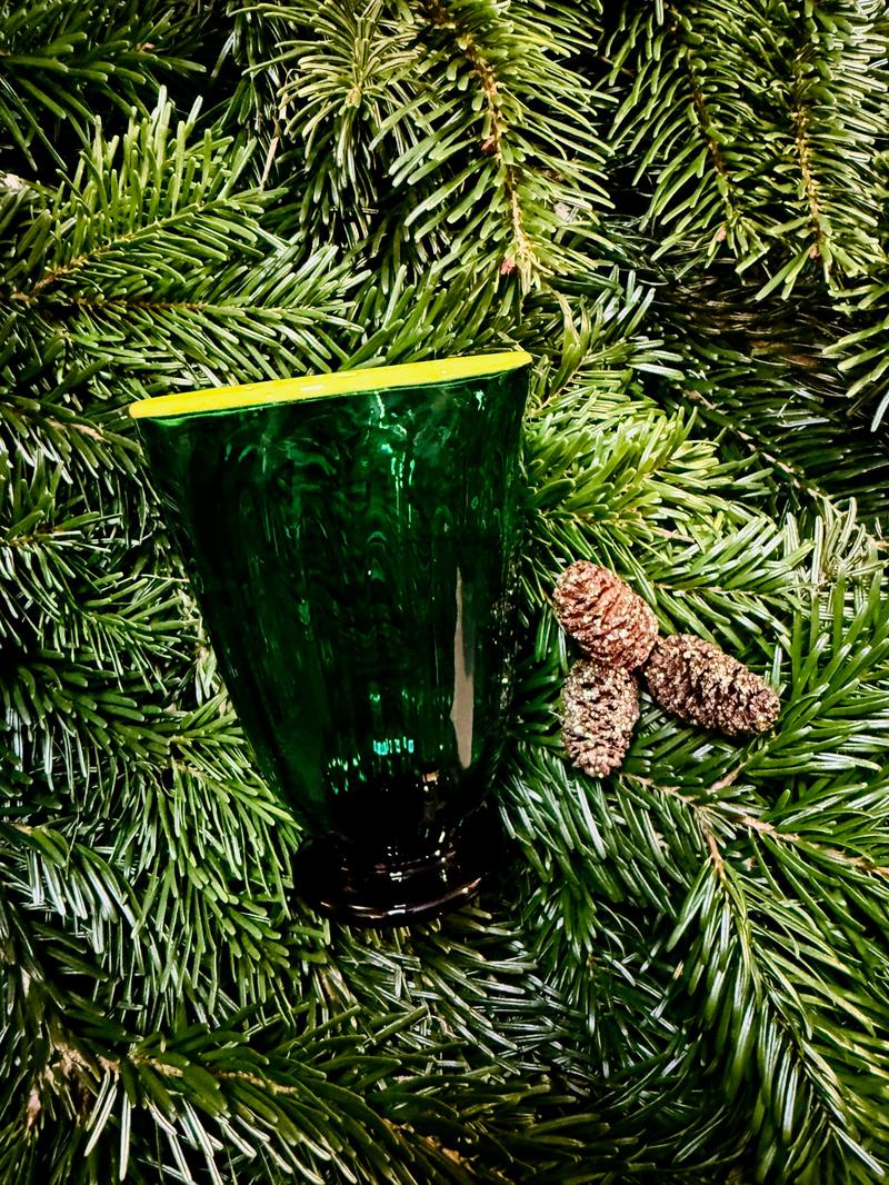 Ein grünes, handgeblasenes Glas mit gelbem Rand steht zwischen dichten Tannenzweigen, daneben liegen mehrere kleine Zapfen. BU