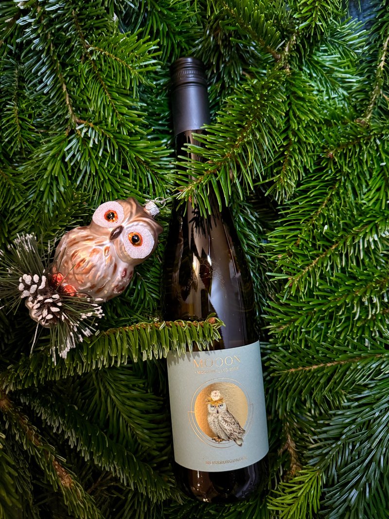 Eine Flasche MOOON-Weißburgunder liegt zwischen frischen Tannenzweigen, daneben hängt eine kleine Eulen-Weihnachtskugel mit glitzernden Details.