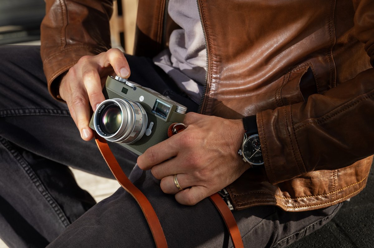 Eine Person hält eine olivgrüne Leica M11-P Safari in beiden Händen. Die Kamera wird im Sitzen gezeigt, kombiniert mit einer braunen Lederjacke und einem cognacfarbenen Kameragurt.