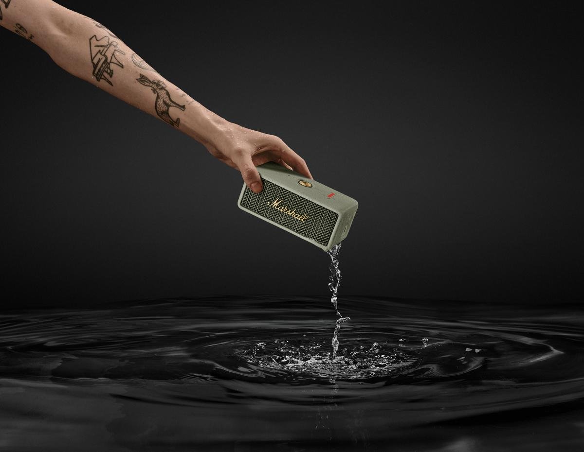 Eine tätowierte Hand hält einen Marshall Emberton III Lautsprecher über eine Wasseroberfläche, während Wasser aus dem Gerät herabfließt.