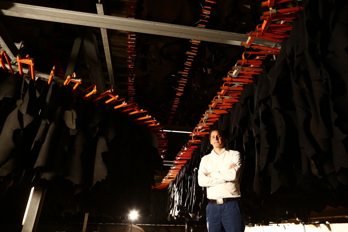 Ein Mann, blaue Hose, weißes Hemd (Thomas Heinen) steht mit verschränkten Armen in einer Gerberei unter der sogenannten Hängetrocknung, umgeben von hängenden Lederhäuten.