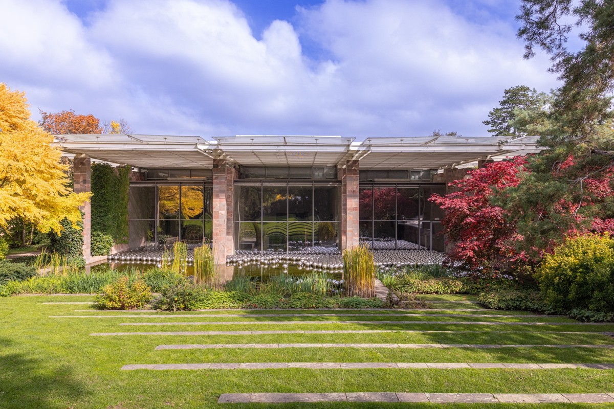 substantial-times-yayoi-kusama-nichts-hat-ein-ende-01 Außenansicht der Fondation Beyeler in Riehen mit der Installation von Yayoi Kusama. Vor der Glasfassade spiegeln sich silberne Kugeln in einem Teich, umgeben von herbstlich gefärbten Bäumen und gepflegtem Rasen.