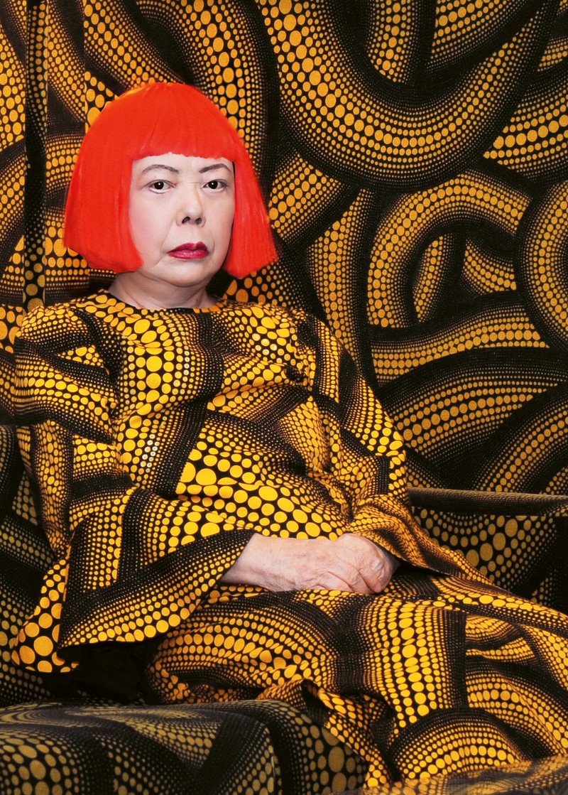 substantial-times-yayoi-kusama-nichts-hat-ein-ende-04 Porträt der Künstlerin, sie sitzt vor einer Wand mit schwarzen Schlangenformen mit gelben Punkten. Ihr Kleid in demselben Muster lässt sie mit dem Hintergrund verschmelzen, nur ihr blass geschminktes Gesicht mit den roten Lippen und der roten Perücke sowie die verschränkten Hände stechen aus dem Bild heraus.