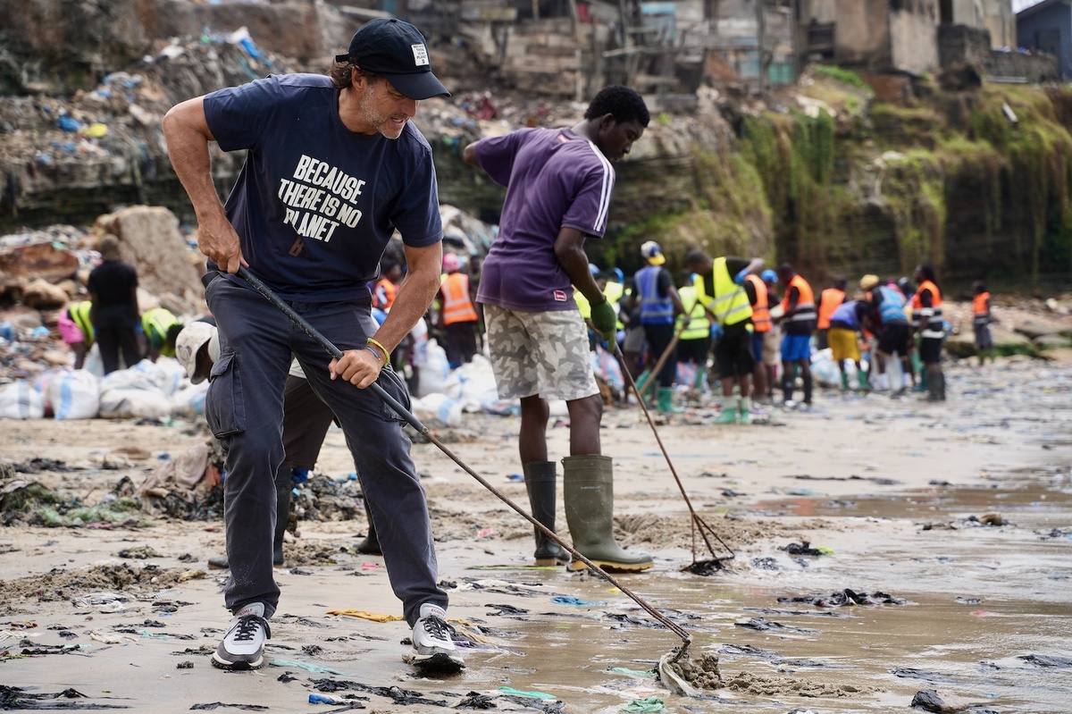 Ein Mann, Javier Goyeneche, und mehrere Helfer harken am verschmutzten Strand von Accra Textilmüll aus dem Sand. Der Mann im Vordergrund arbeitet mit einer Harke und trägt ein T-Shirt mit dem Aufdruck „Because there is no Planet B“.