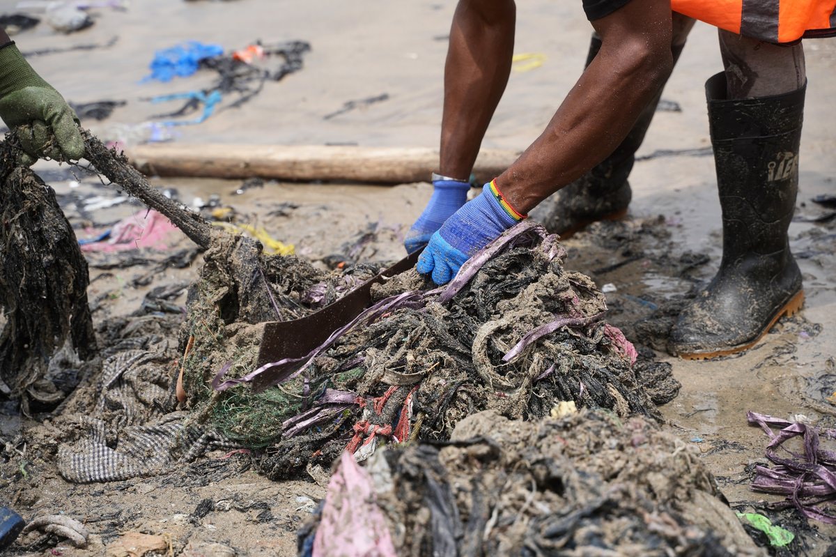 Nahaufnahme von Helfern am Strand, die mit Handschuhen und einer Machete verfilzte Textilabfälle aus dem Sand schneiden und lösen.