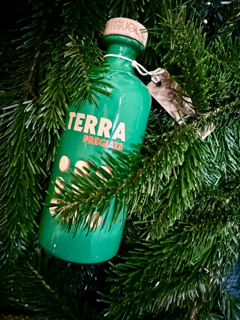 Eine smaragdgrüne Flasche Olivenöl der Sorte „Terra Pregiata“ von Oleificio Guglielmi liegt eingebettet zwischen üppigen, dunkelgrünen Tannenzweigen. Der Korkverschluss trägt eine feine Gravur, und am Flaschenhals hängt ein kleines braunes Etikett aus Papier. Die glänzende Oberfläche der Keramikflasche reflektiert das Licht, während die Tannenzweige teilweise den unteren Teil der Flasche verdecken. Das gesamte Bild vermittelt eine natürliche, festliche Atmosphäre.
