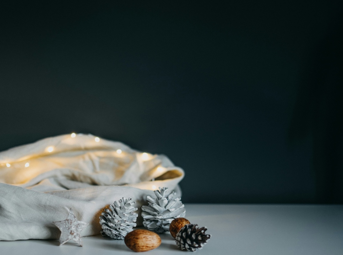 Weihnachtliches Stillleben mit hellem Leinensack, warmweißer Lichterkette, weißen Tannenzapfen und Nüssen auf einem Tisch vor dunklem Hintergrund.