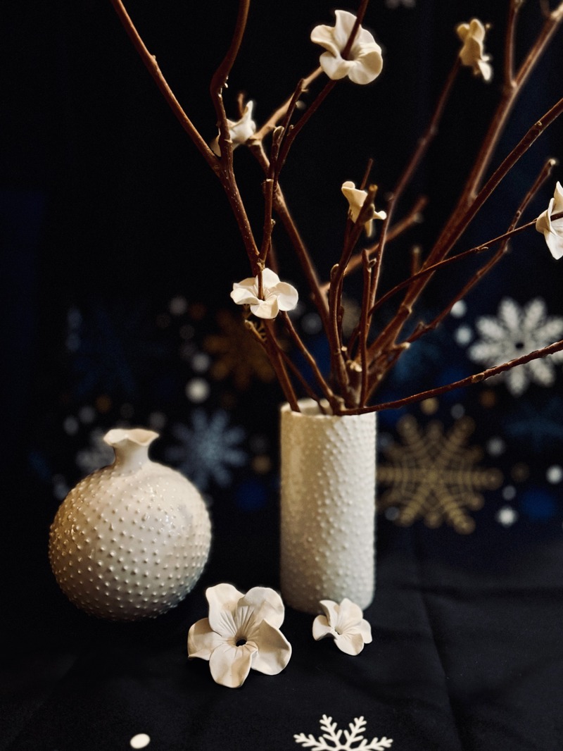 Keramikvasen und Porzellanblüten von Coco Loves Clay: eine kugelförmige und eine zylindrische weiße Vase mit strukturierter Oberfläche, dazu unglasierte Blüten und Zweige, arrangiert auf dunklem Untergrund mit weihnachtlichen Motiven.
