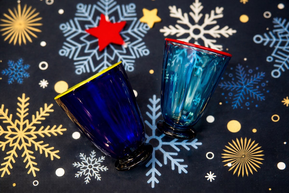 Zwei handgefertigte Trinkgläser aus blauem Glas mit farbig abgesetzten Rändern, arrangiert auf dunkelblauem Stoff mit winterlichem Schneeflocken- und Sternmuster.