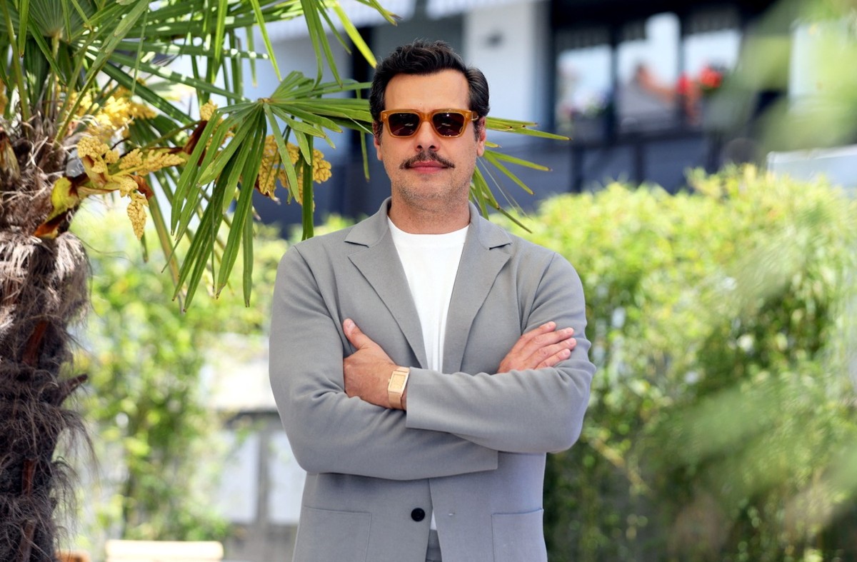 Ein Mann mit Sonnenbrille steht im Freien vor grüner Vegetation und Palmen, die Arme vor der Brust verschränkt; an seinem Handgelenk trägt er eine rechteckige Uhr mit Rotgoldgehäuse und Milanaise-Armband.