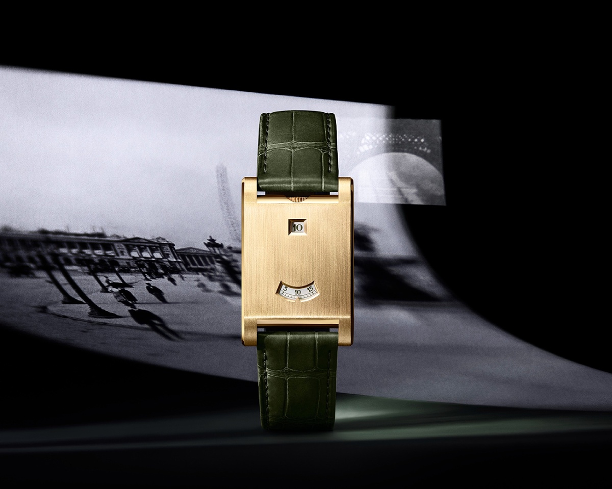 Rechteckige Cartier-Uhr in Gelbgold mit digitaler Stunden- und Minutenanzeige, ausgeschnitten aus einem goldenen geschlossenen Gehäuse, präsentiert vor einer schwarz-weißen, unscharf projizierten historischen Szenerie; das Modell ist an einem tannengrünen Lederband befestigt.