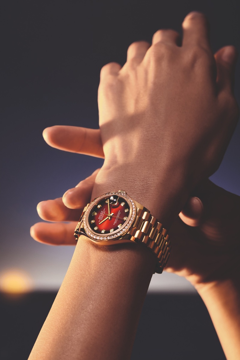Weibliches Handgelenk mit goldener Rolex-Armbanduhr; die Datejust 31 besitzt ein rot-ombré-Zifferblatt mit Diamantindizes und diamantbesetzter Lünette, getragen an einem President-Band.
