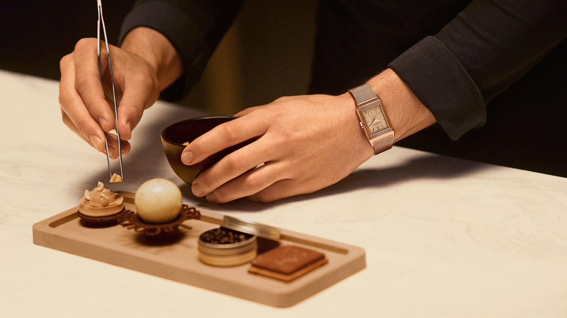 Detailfoto: Hände eines Chocolatiers, der mit einer Pinzette kunstvolle Schokoladenkreationen auf einem Holztablett arrangiert; am Handgelenk trägt er eine Jaeger-LeCoultre Reverso in Rotgold mit Milanaise-Armband.